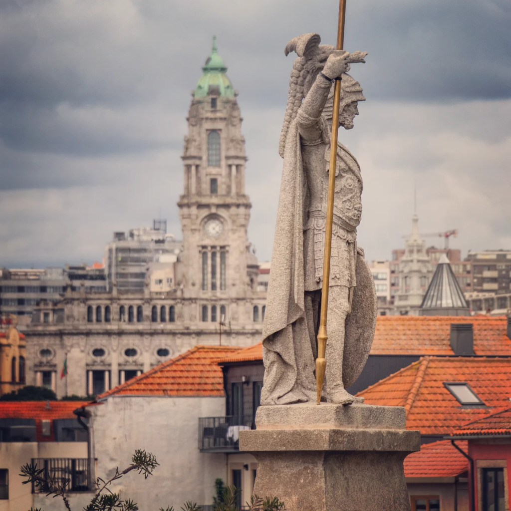 Close-up of a stone statue of a Roman soldier holding a spear, with Porto’s City Hall clock tower and modern buildings blurred in the background.