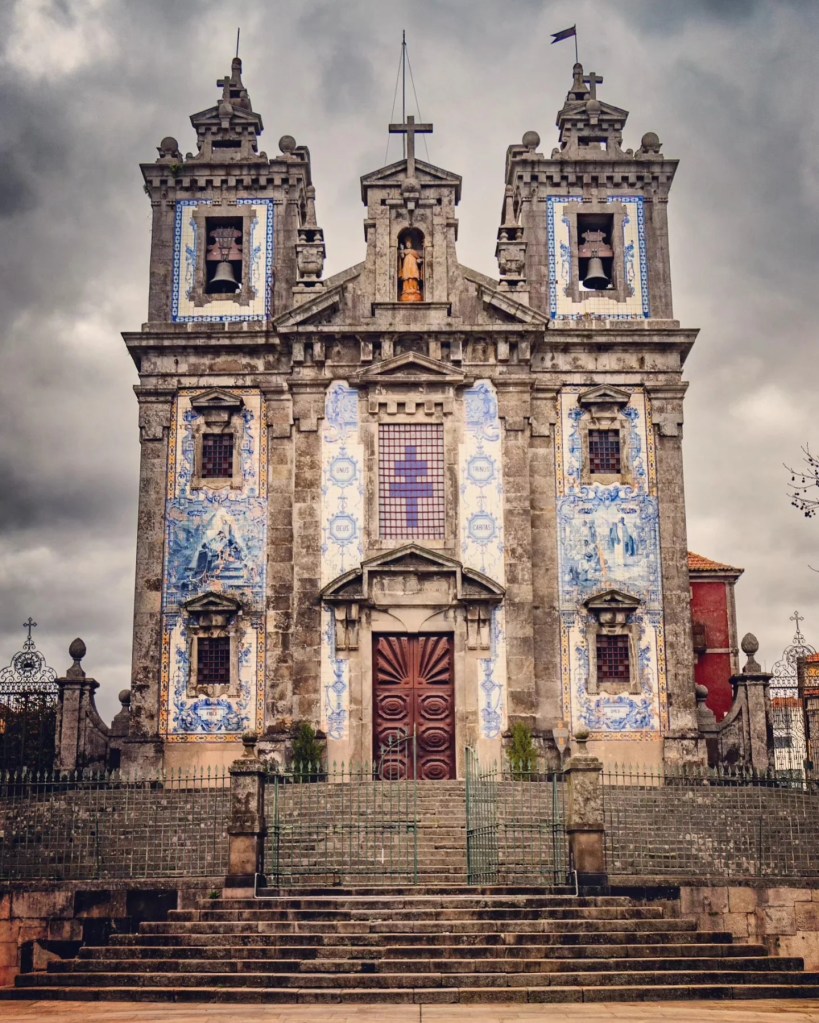 Front façade of the Igreja de Santo Ildefonso in Porto, Portugal, featuring blue and white azulejo tiles under moody clouds.