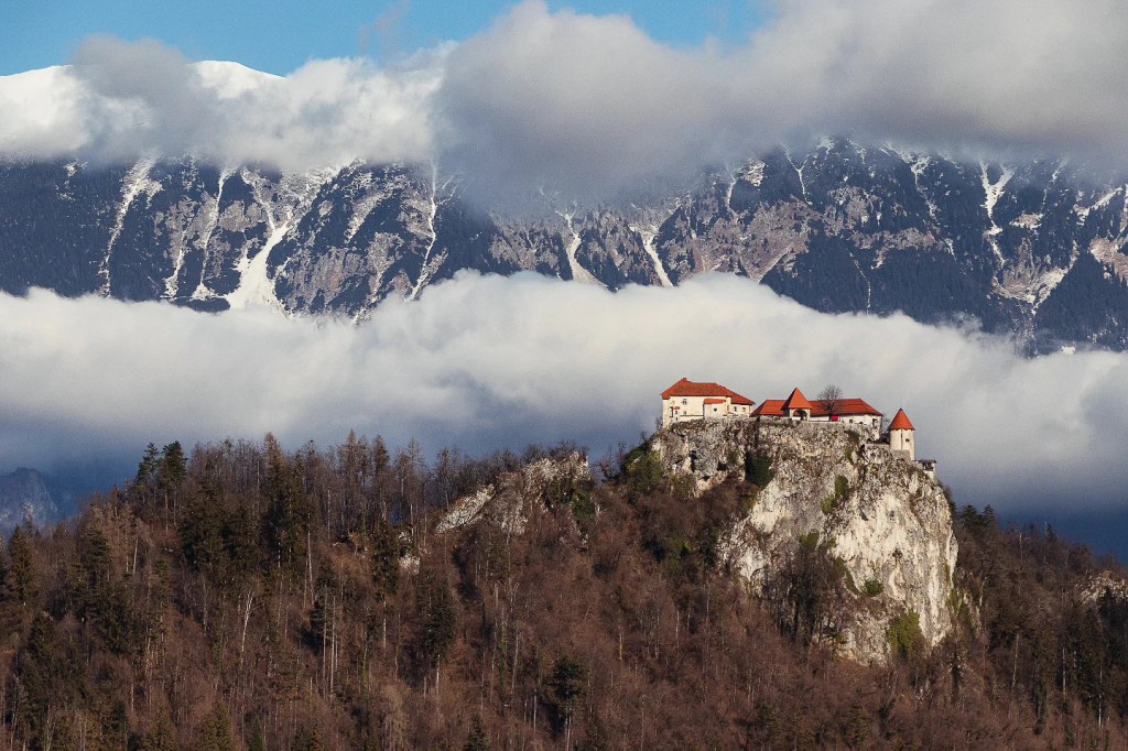 Bled Castle perched on a rocky cliff surrounded by winter trees, with the dramatic Julian Alps towering behind, partially shrouded in clouds.