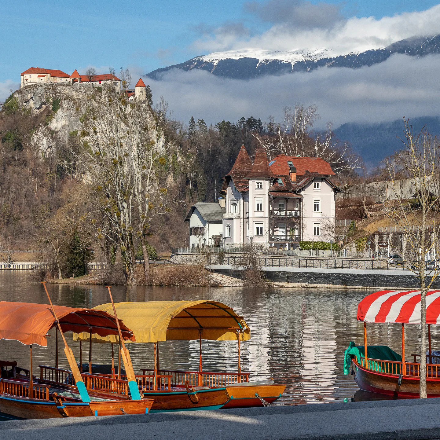 Traditional Pletna boats with colorful canopies docked on Lake Bled, with a view of lakeside villas and Bled Castle on the cliff in the background.