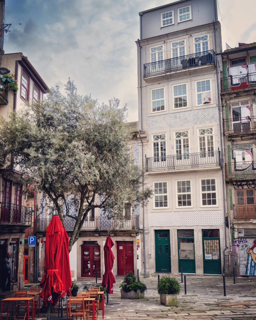 A quiet square with outdoor tables and bright red closed umbrellas, surrounded by tall, narrow buildings with tiled facades in Porto.