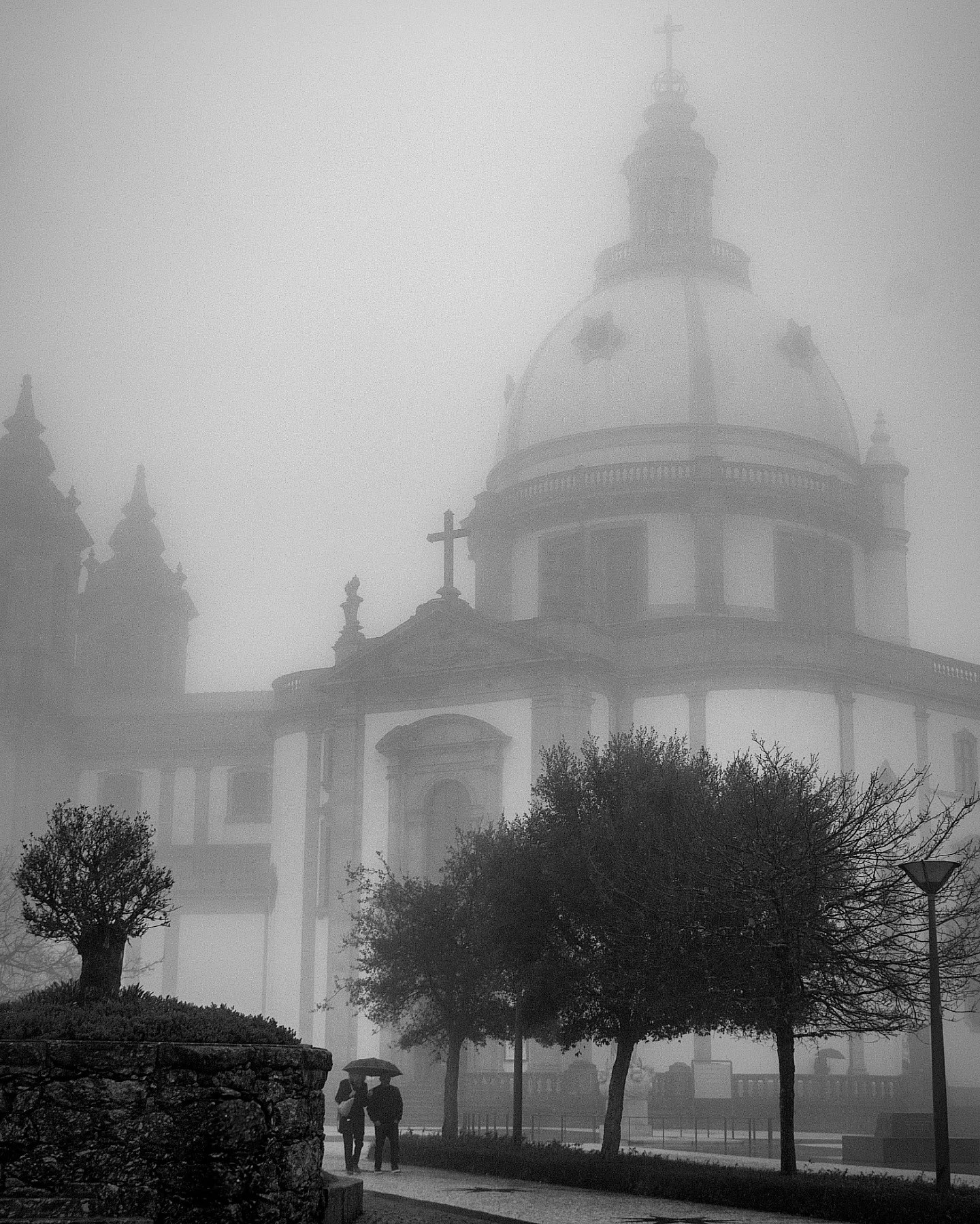 The large dome of The Sanctuary of Our Lady of Sameiro looms ghostlike through thick morning fog, with two people walking under umbrellas in the foreground.