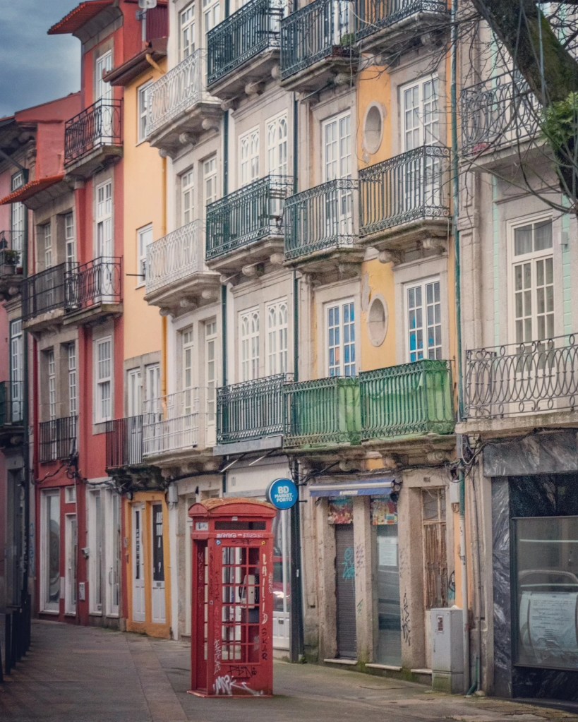 A red British-style phone booth stands in front of a row of colorful, weathered apartment buildings with wrought iron balconies in Porto.