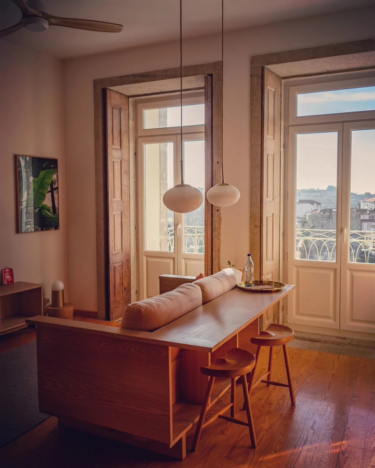 Cozy and sunlit interior of an apartment in Porto, featuring tall wooden shutters, soft pendant lighting, and a simple wooden dining bar with stools in front of the view.