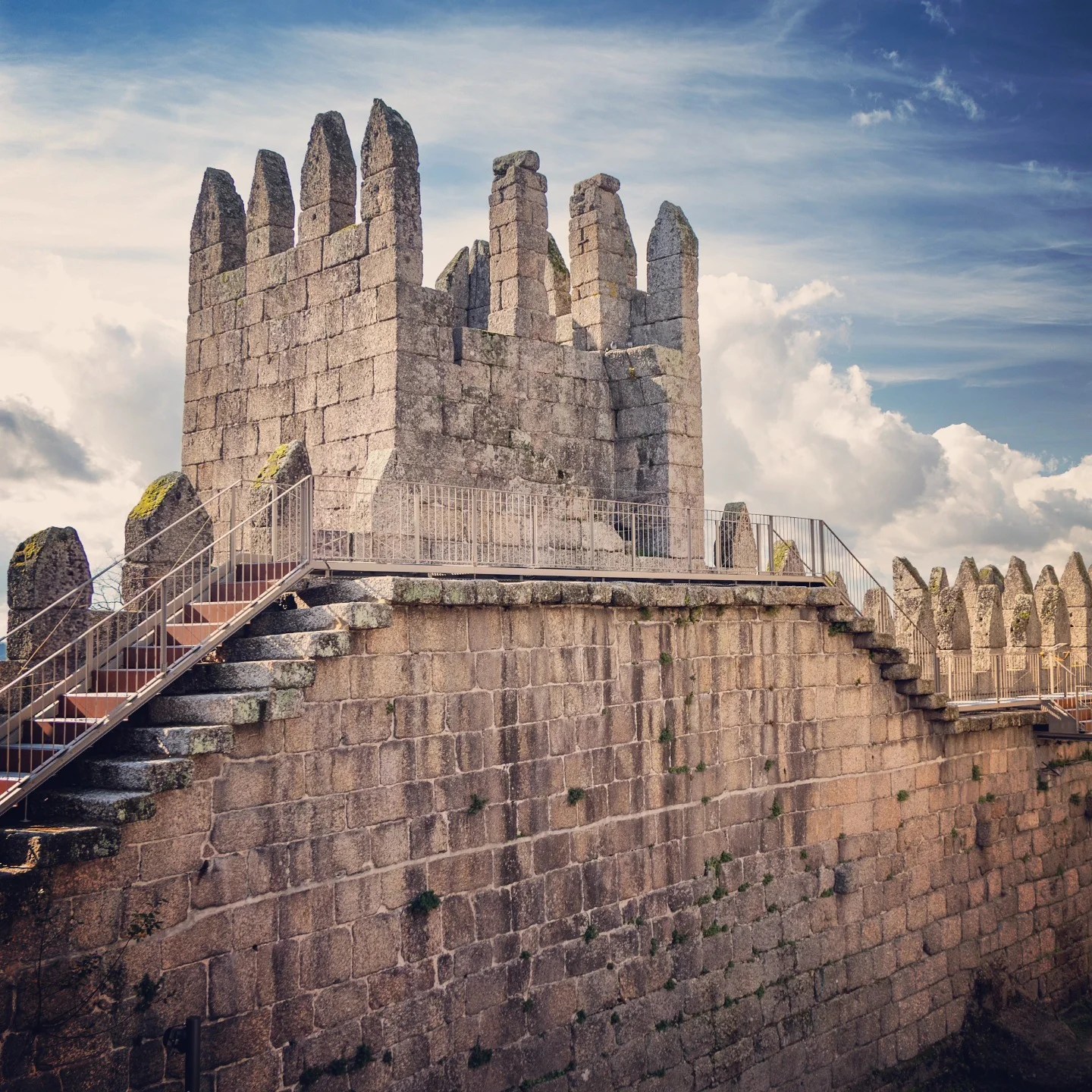 A section of the crenellated stone walls and battlements of Guimarães Castle under a partly cloudy sky, with modern metal stairs providing visitor access.