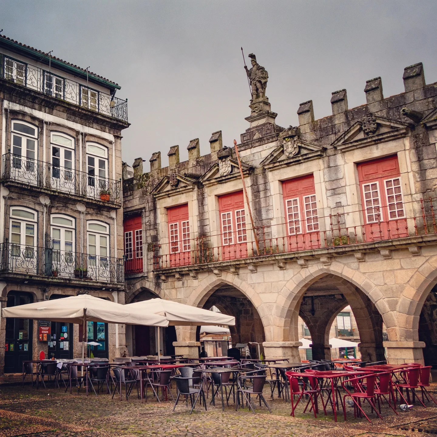 The arched arcade and red-shuttered windows of the old council chambers (Antiga Câmara Municipal) in Largo da Oliveira, Guimarães, with empty outdoor café seating below.