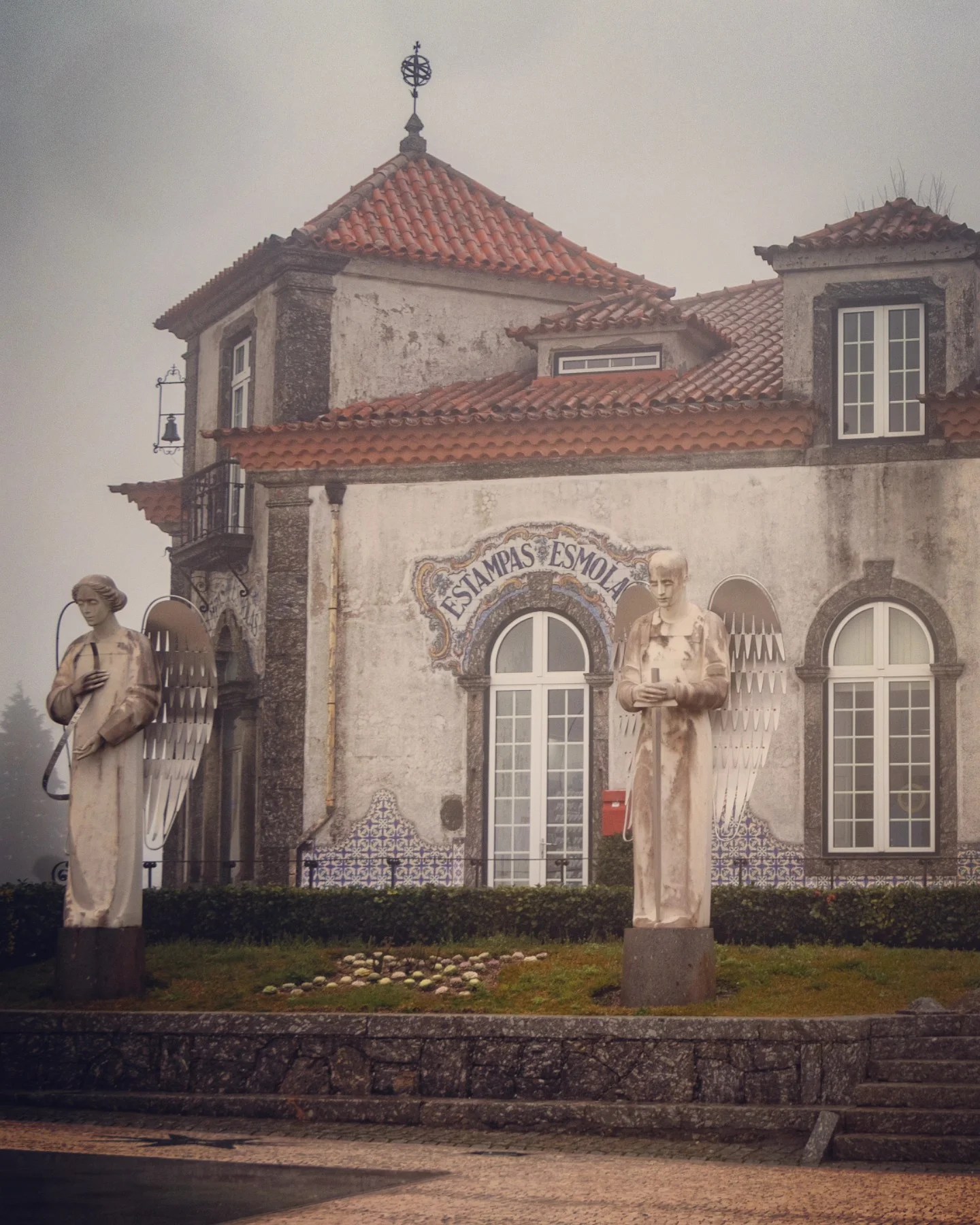 Two angelic statues stand in front of the Casa das Estampas shop at The Sanctuary of Our Lady of Sameiro. The whitewashed building features blue tile accents and a terracotta roof.