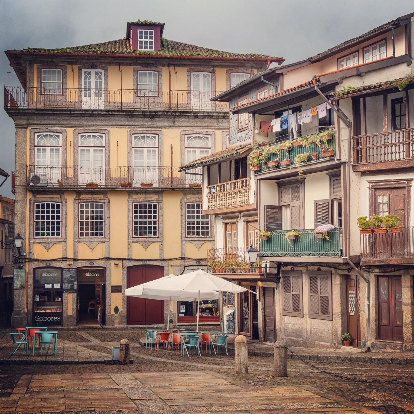 Colorful chairs and white parasols at an outdoor café on Largo da Oliveira square in Guimarães, surrounded by historic stone and stucco buildings with wrought-iron balconies.