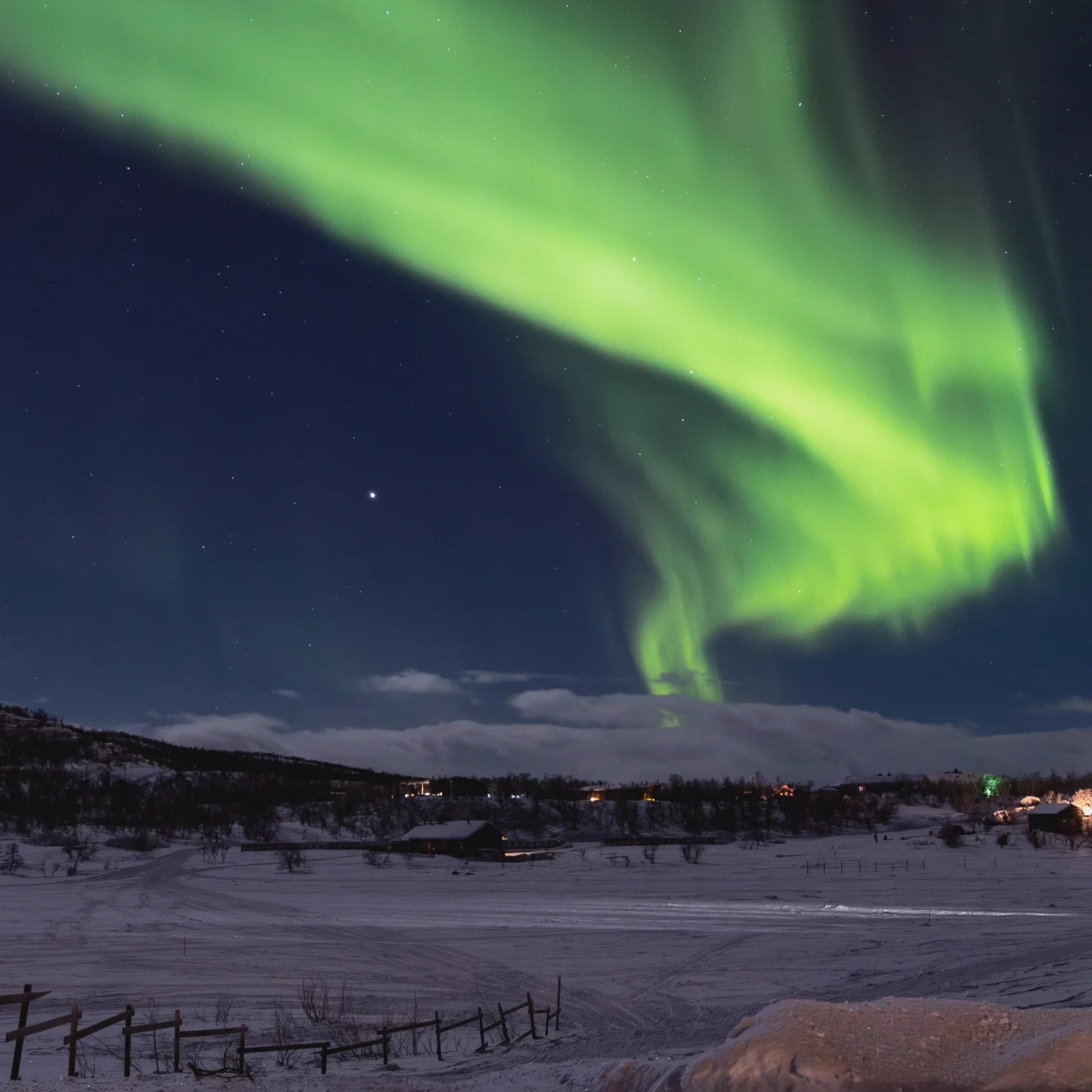 Bright green Northern Lights swirl over a snowy Finnish landscape with scattered cabins and stars above.