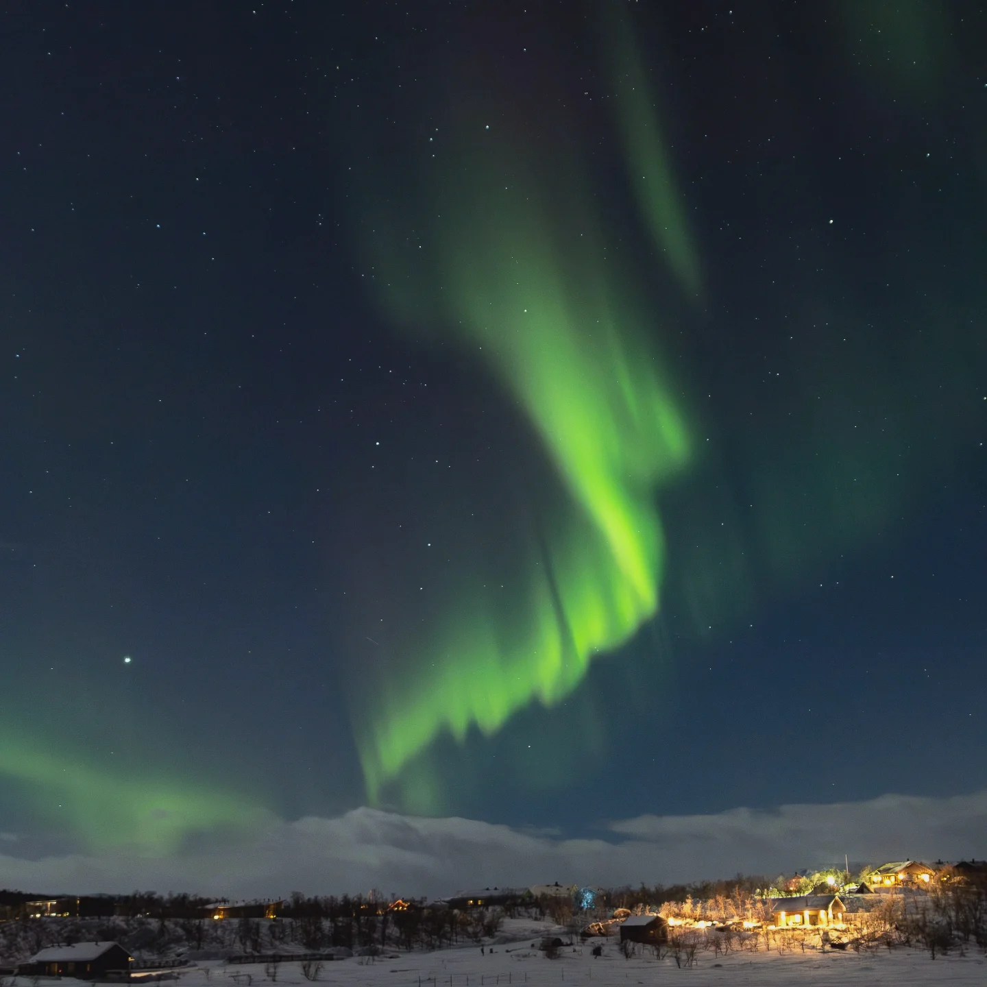 Intense green Northern Lights streak across the sky above a glowing village nestled in snowy hills.