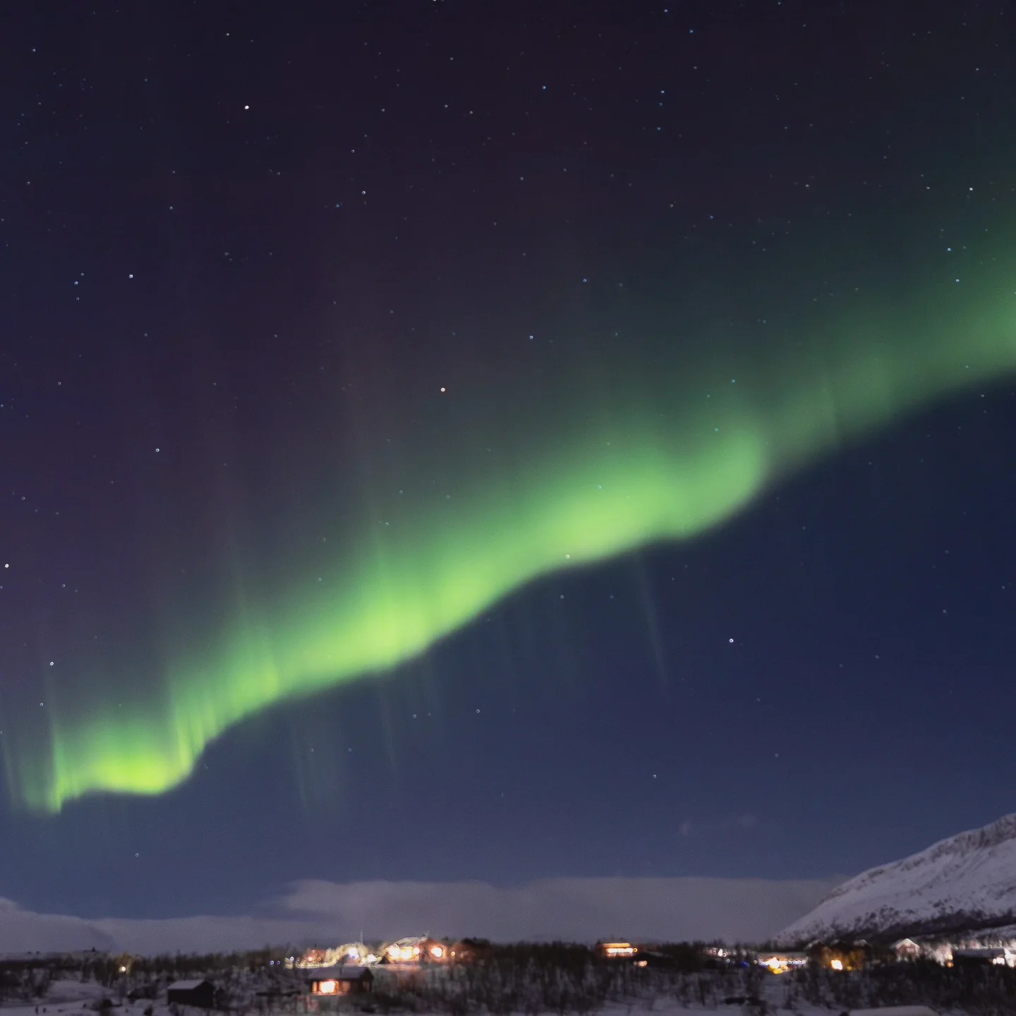 A wide curtain of Northern Lights illuminates the night sky over a snowy town and surrounding hills.