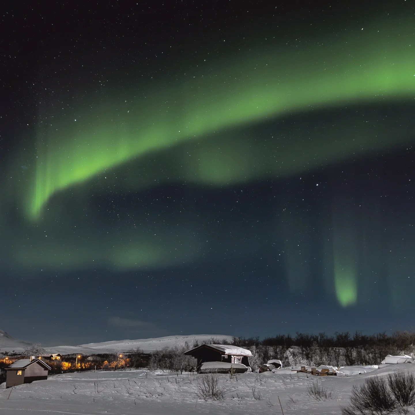 A glowing arc of Northern Lights over a snowy Finnish cabin, stars twinkling above a wintry scene.