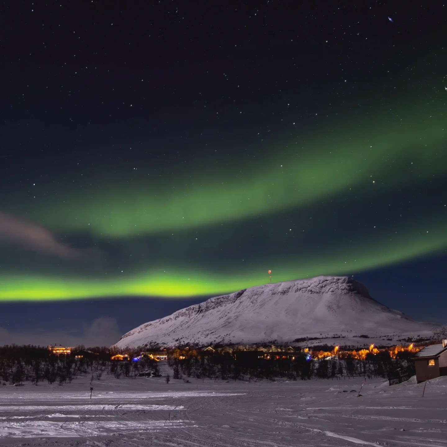 Green Northern Lights arch over a snow-covered mountain and village in Finland, with stars filling the night sky.