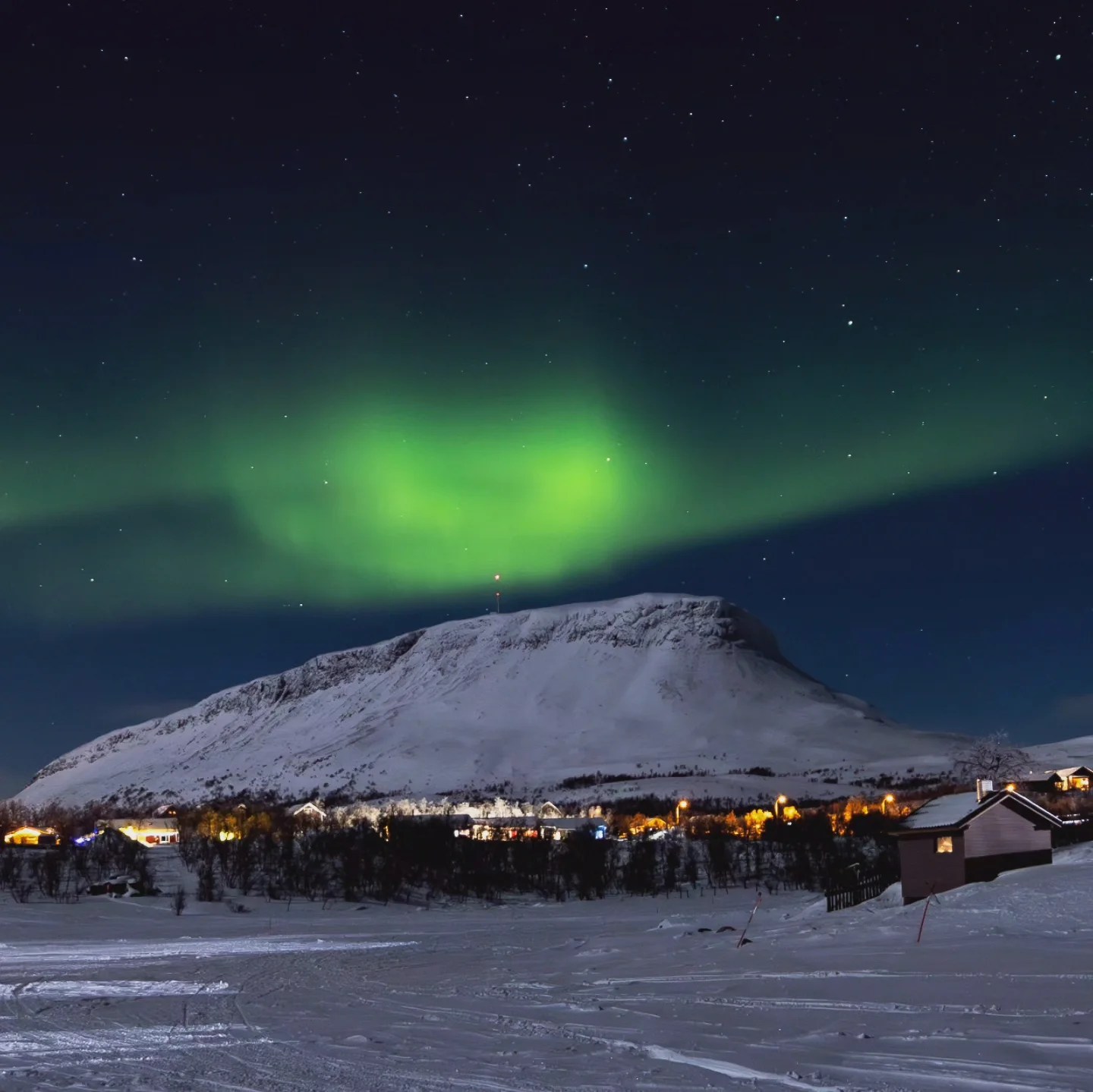 Bright green aurora hovers above a snow-dusted peak and glowing village lights during a clear Arctic night.