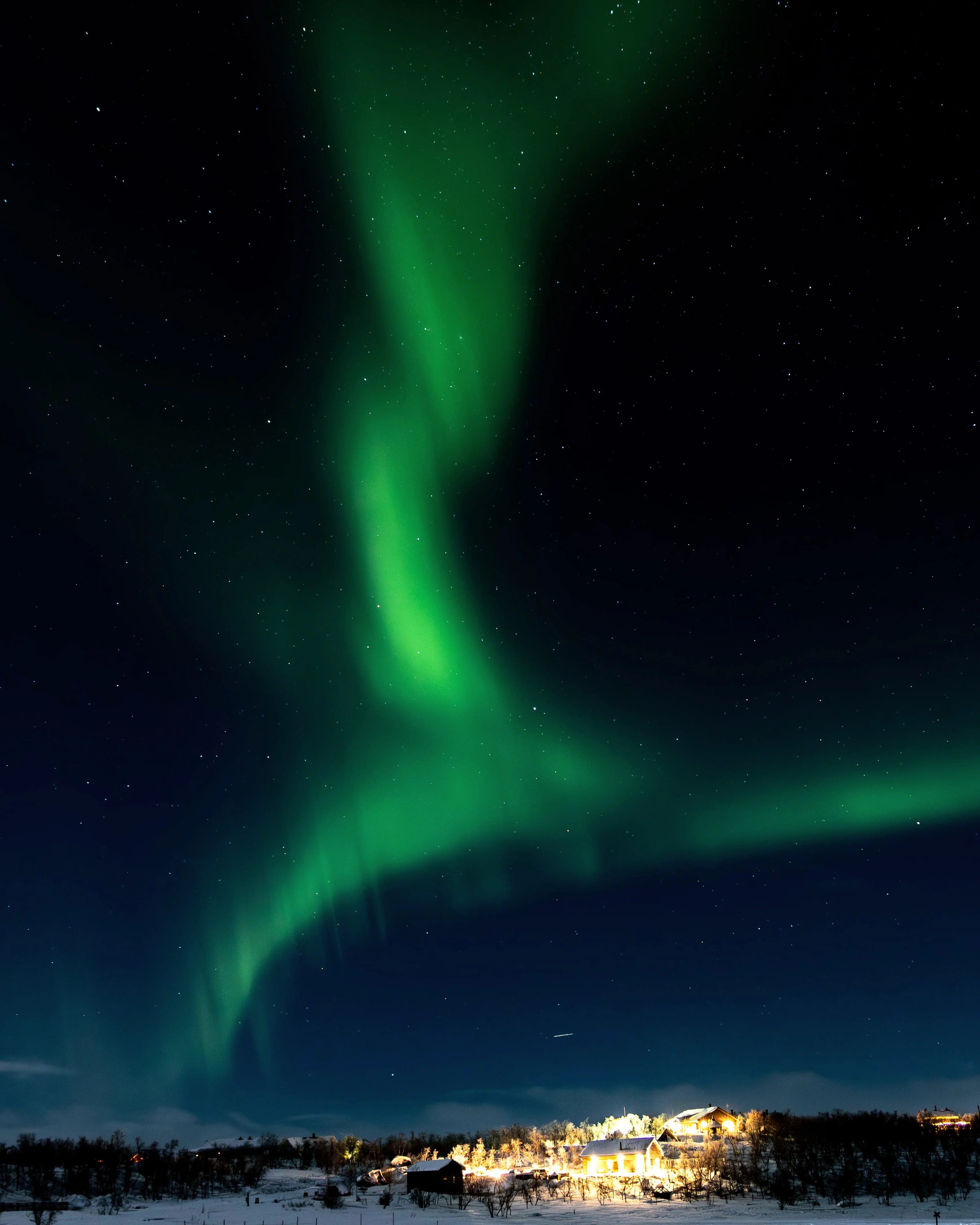 Green aurora lights flow down from the sky above a warmly lit Finnish village under the stars.