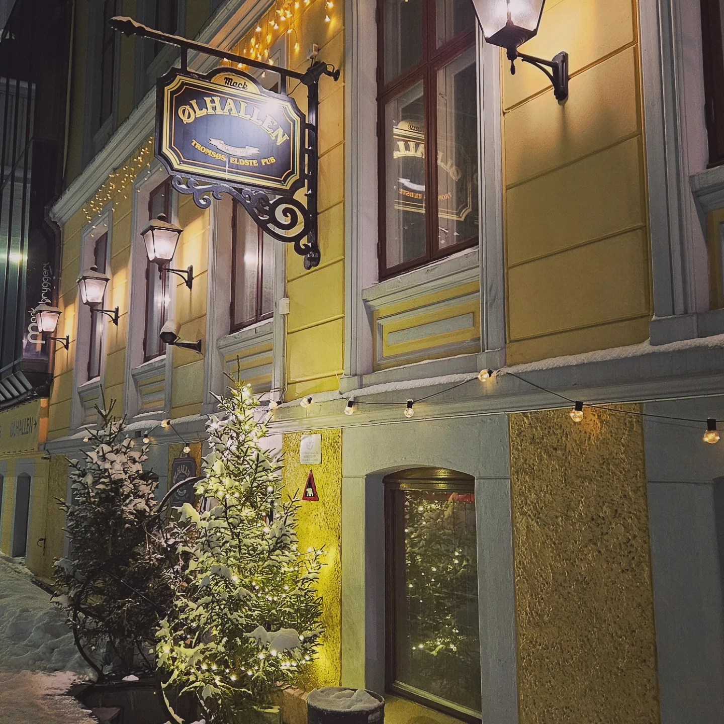 The warmly lit yellow exterior of Ølhallen pub at night, with snow-covered trees, string lights, and a decorative sign.