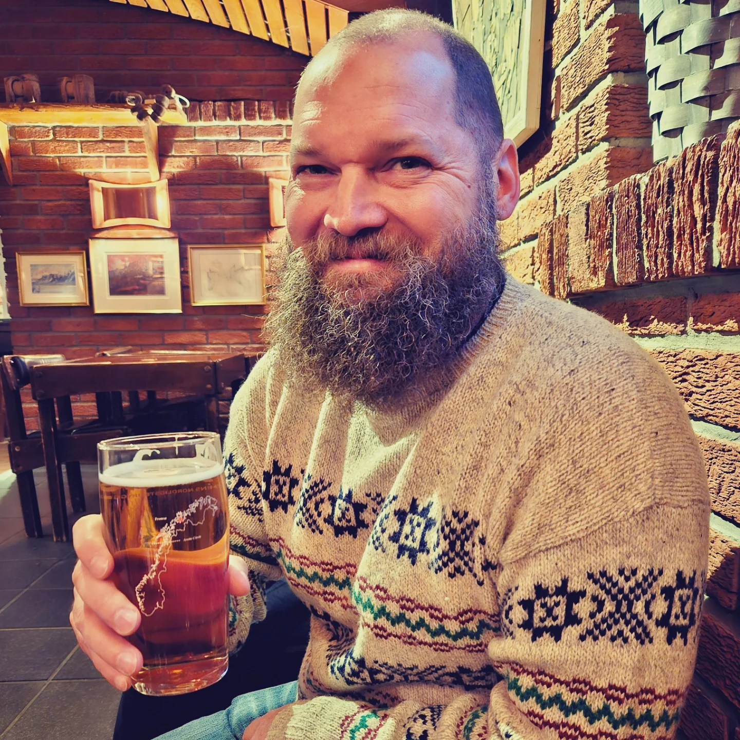 A bearded man in a Nordic sweater smiles while holding a pint of beer inside Ølhallen’s cozy brick interior.