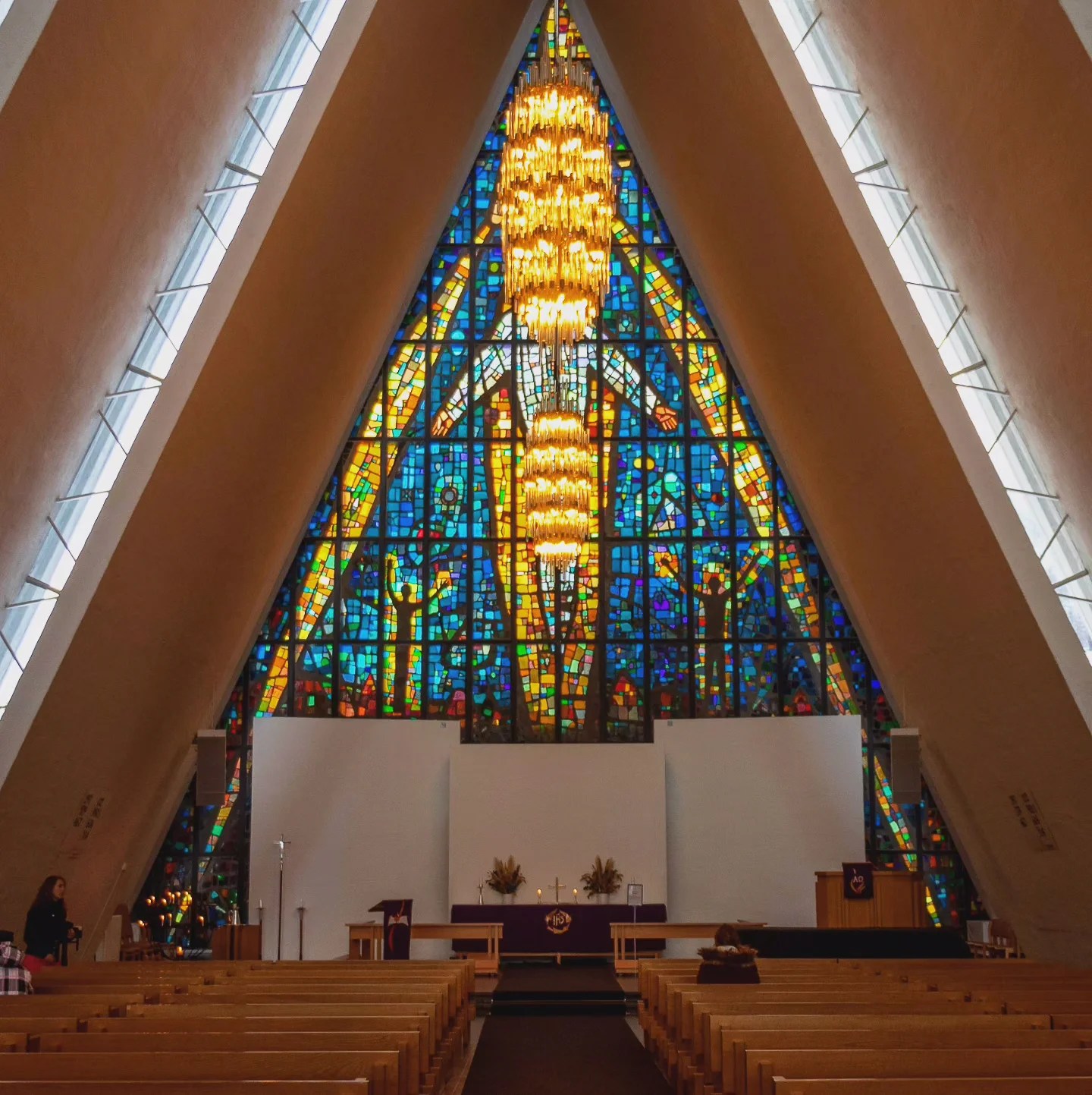 Interior of the Arctic Cathedral showing rows of wooden pews and a brilliant stained glass window with a large chandelier.