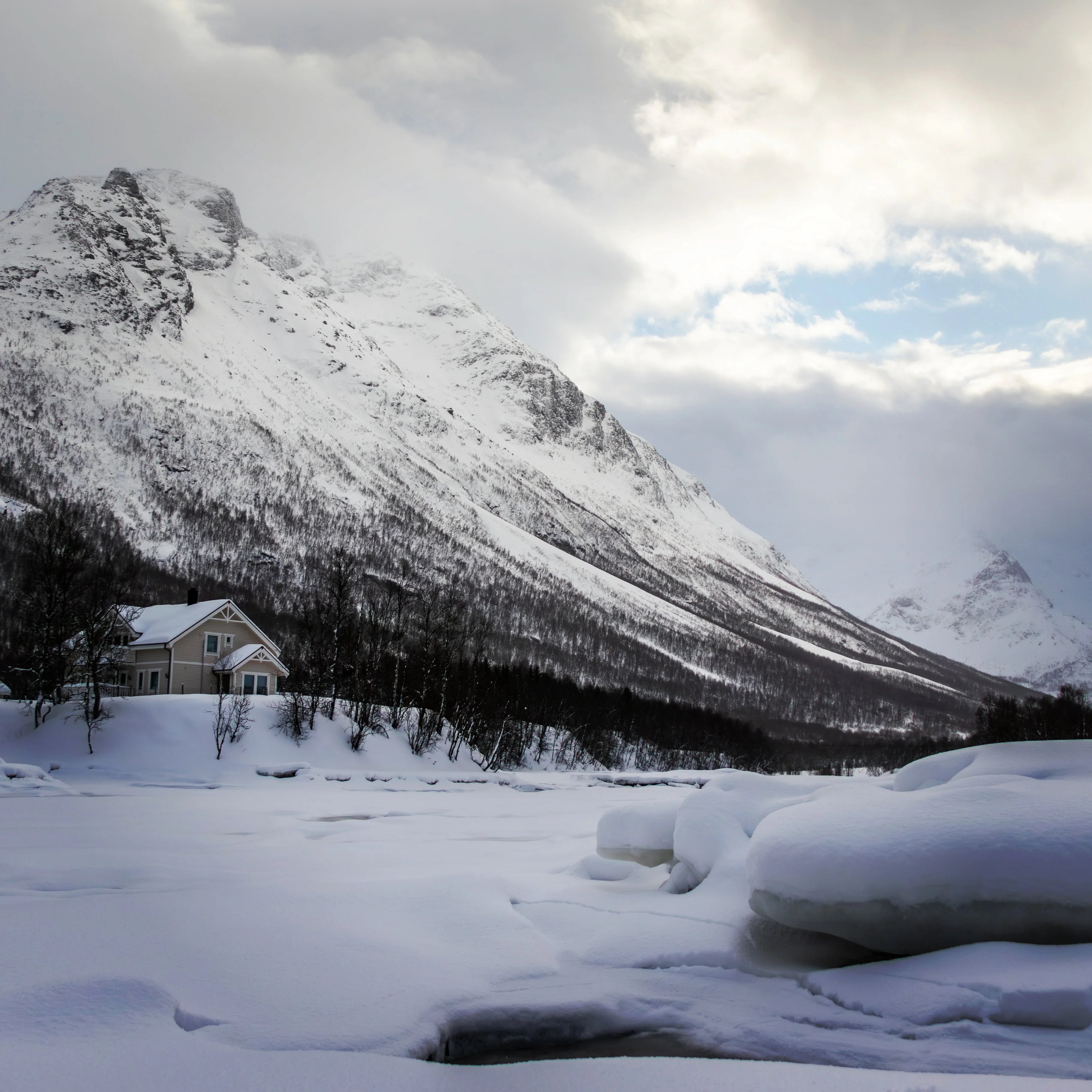 A cozy house sits near snowdrifts beneath a dramatic mountain in the Tromsø countryside, under a break in the clouds.