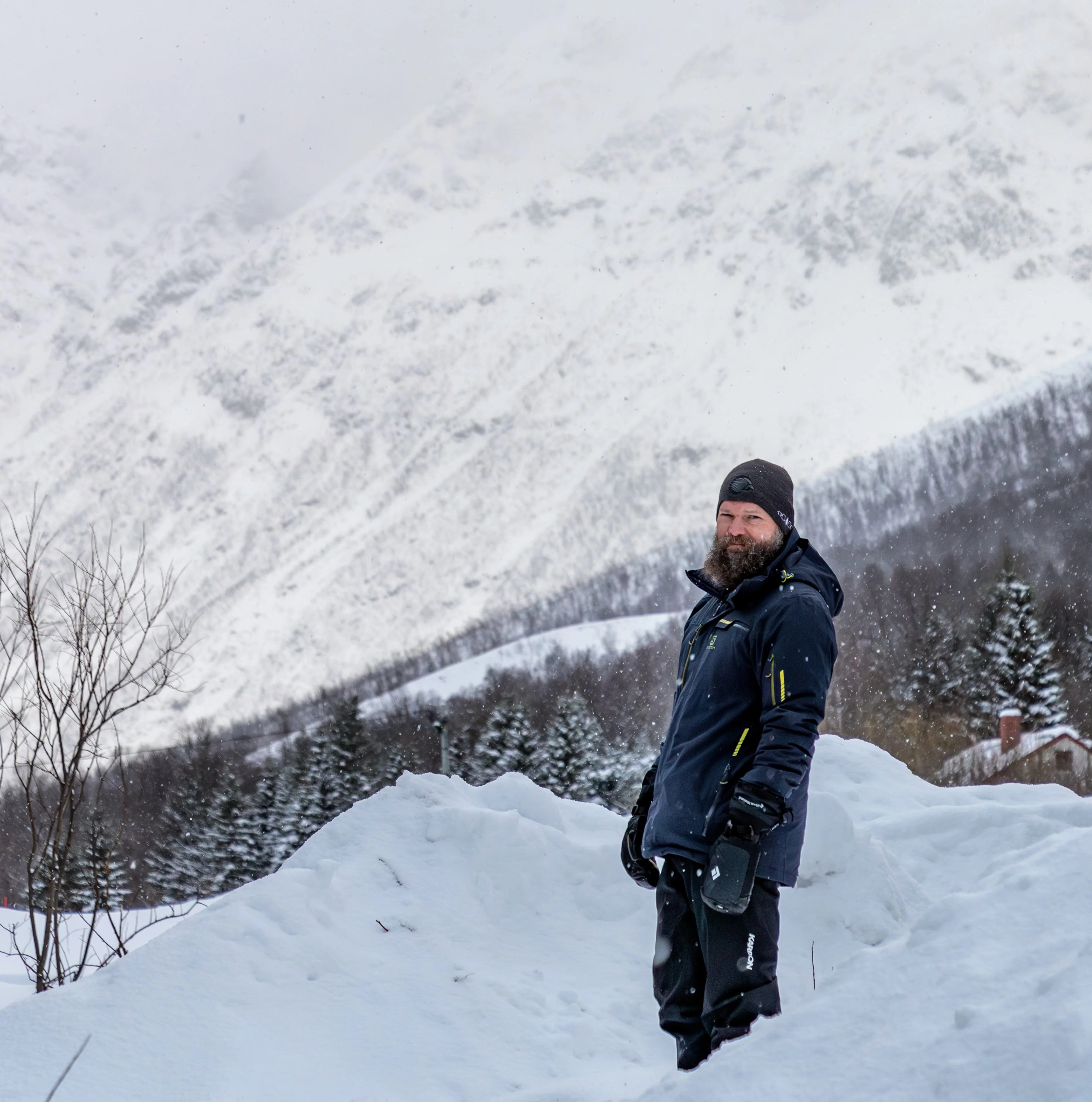 A bearded man in winter gear stands knee-deep in snow, with a steep, forested mountain ridge rising behind him.