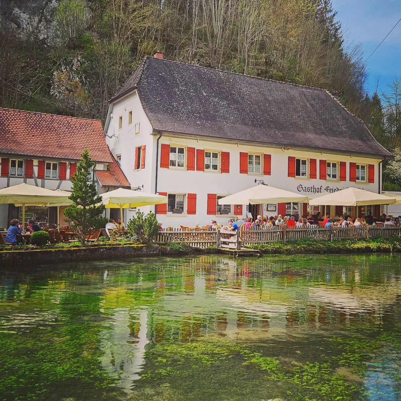 Outdoor dining area of Gasthof Friedrichshöhle near Wimsen Cave, with people enjoying food and drinks under white umbrellas next to the river.
