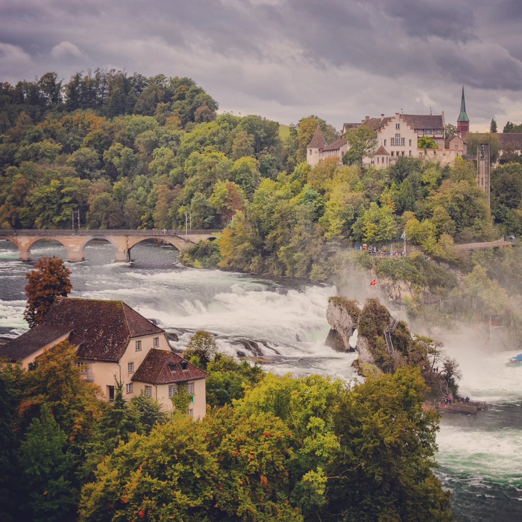 Panoramic view of Rhein Falls with Schloss Laufen perched above roaring white water under brooding clouds.