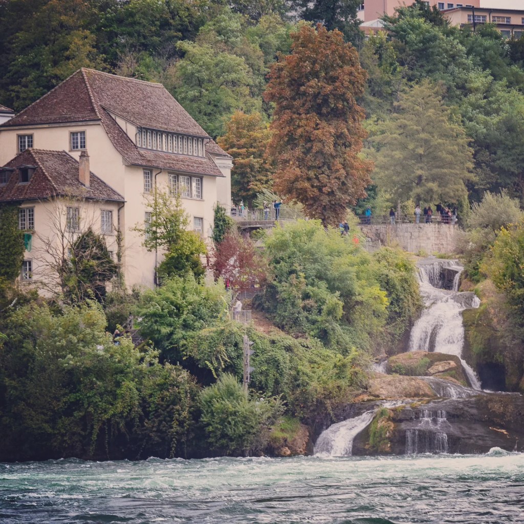 Riverside villa framed by mixed-season foliage beside a smaller cascade near Rhine Falls.