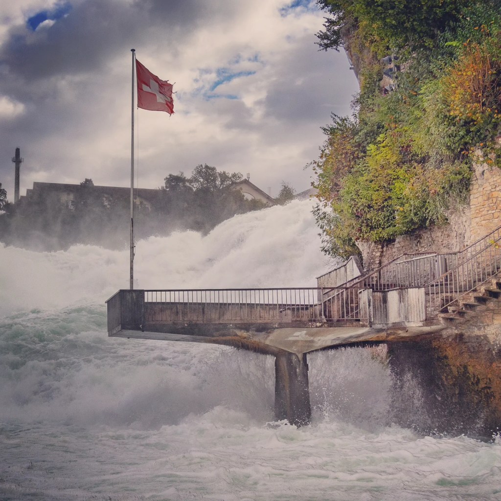 Swiss flag flying above a misty viewing platform at the base of Rhine Falls’ thundering water.