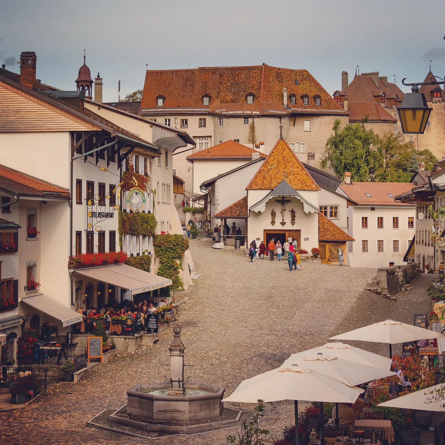 Cobblestoned main square of Gruyères lined with flower-decked chalets, fountain and chapel.