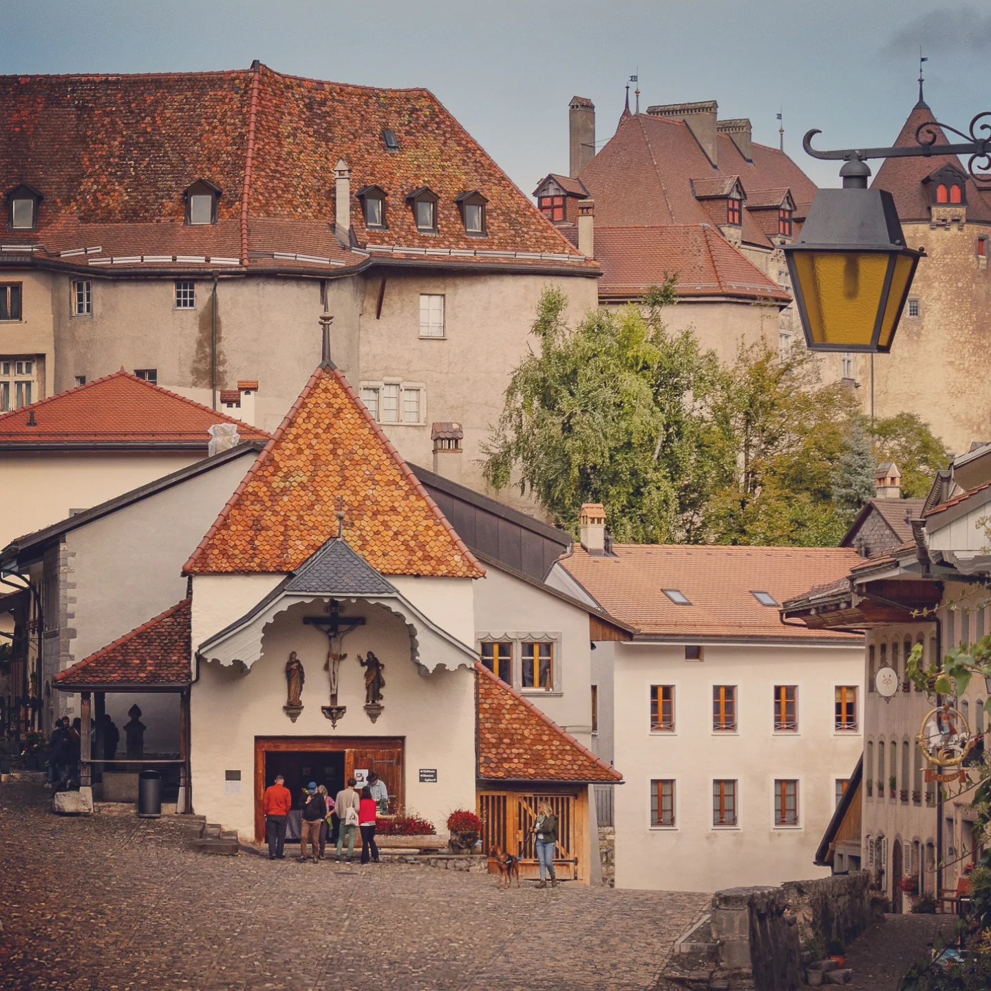 Cobblestoned square with a small chapel topped by a terracotta roof; medieval castle walls and turrets rise behind.