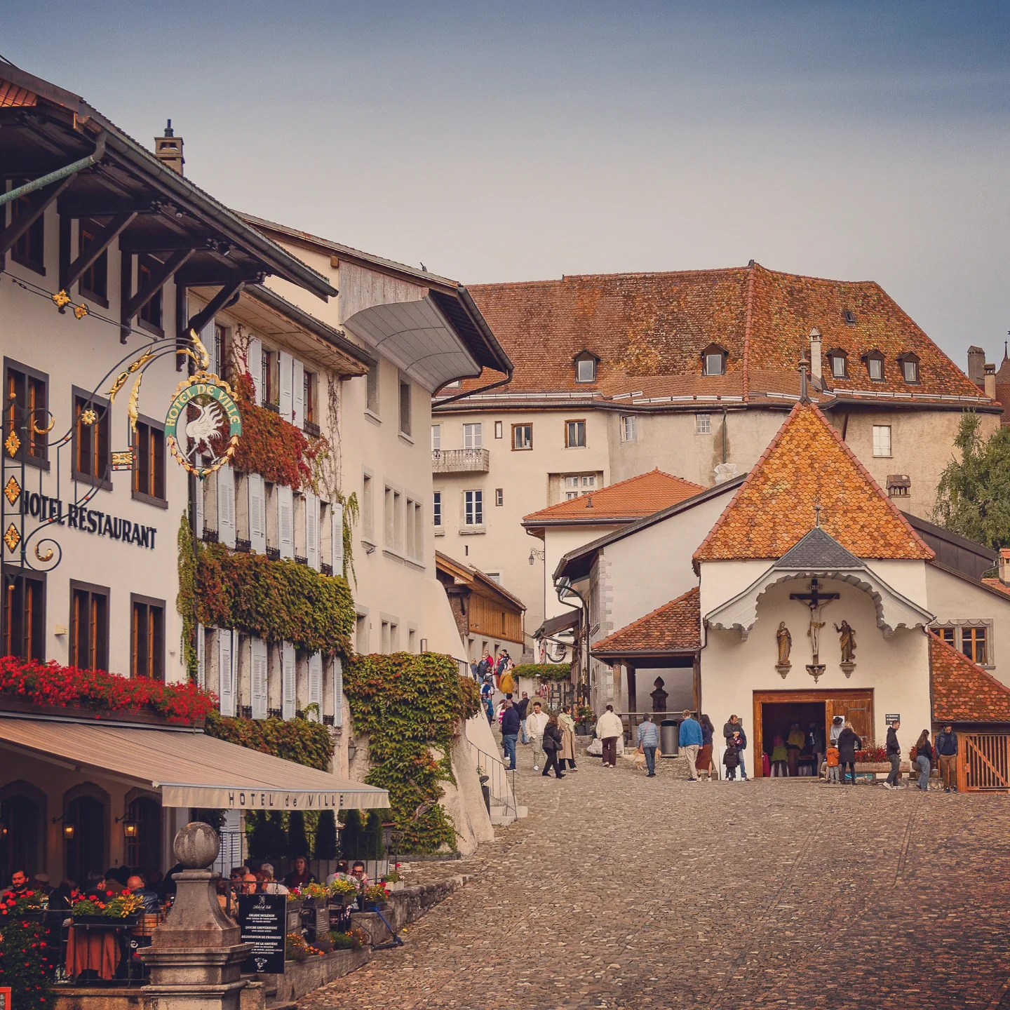 Gruyères’ main street with ivy-clad Hôtel de Ville restaurant on the left and chapel at centre; visitors stroll the cobbles.
