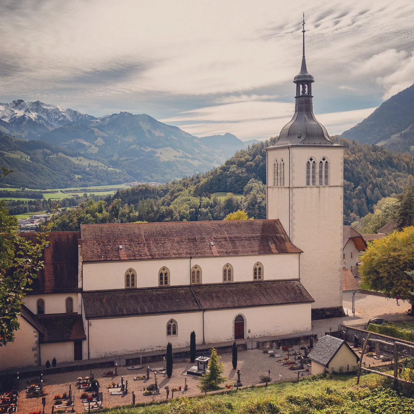 Church of Saint-Théodule in Gruyères—white nave and tall slate spire—overlooking a wide green valley with snow-dusted Pre-Alps behind.