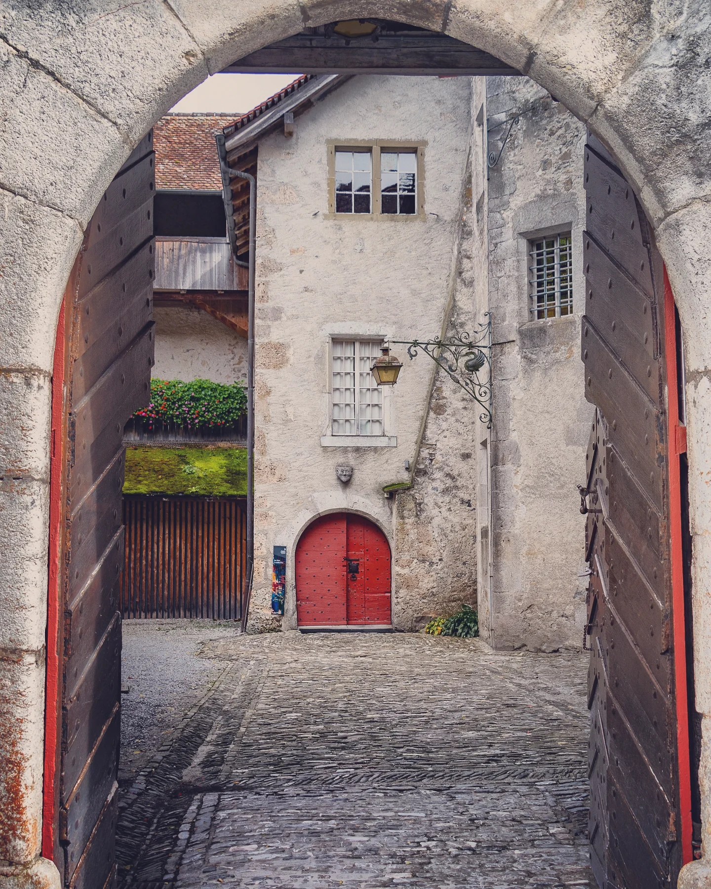 View through a stone archway into the cobbled castle courtyard of Château de Gruyères with a bright red arched door as an entrance into the castle.