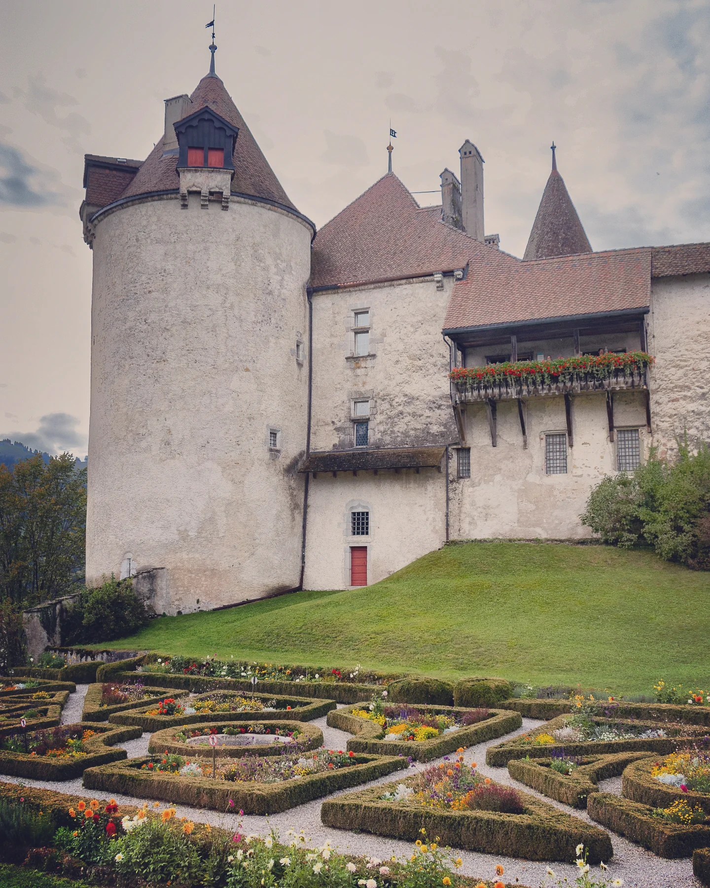 Cylindrical tower and walls of Gruyères Castle rising above a formal knot garden of box hedges and flowers.