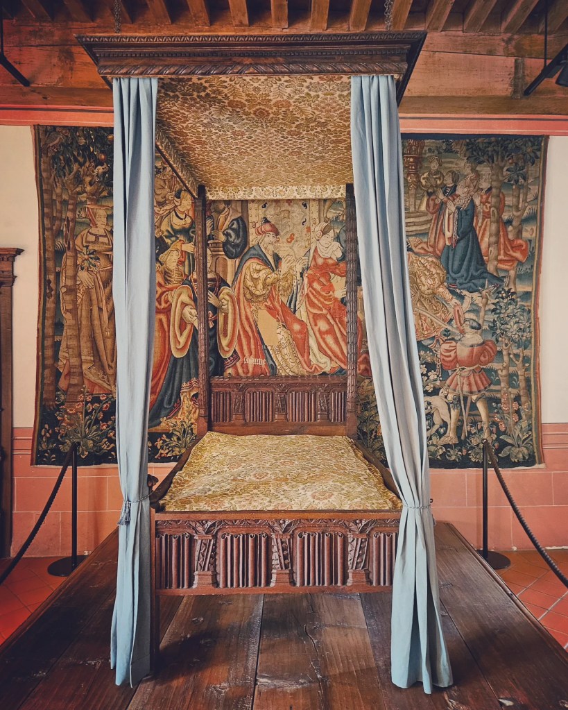 Ornate four-poster bed with pale blue curtains and rich medieval tapestries in a castle bedroom.