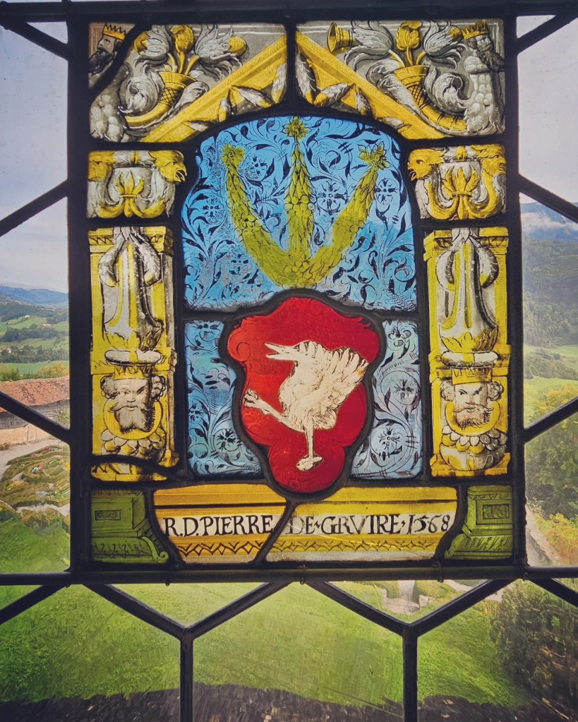 Close-up of stained-glass pane (1568) showing Gruyères’ crowned crane emblem with alpine scenery visible through the window.
