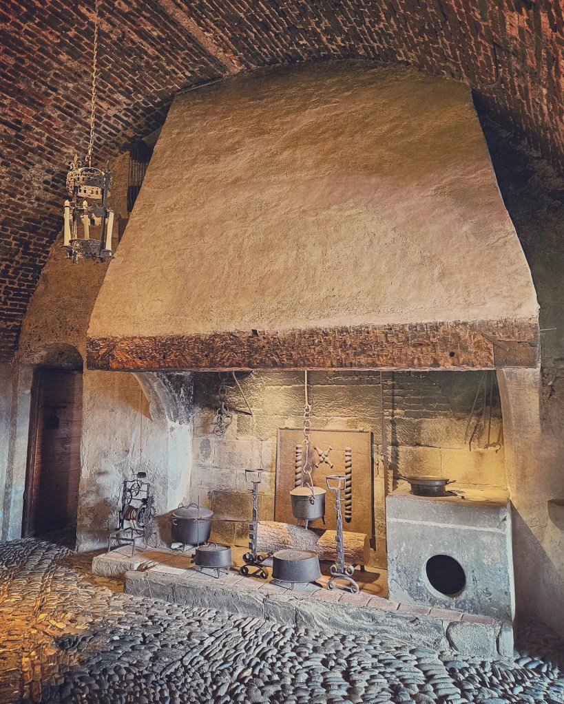 Vaulted brick kitchen at Château de Gruyères with a massive hearth, iron cauldrons and cobbled floor.