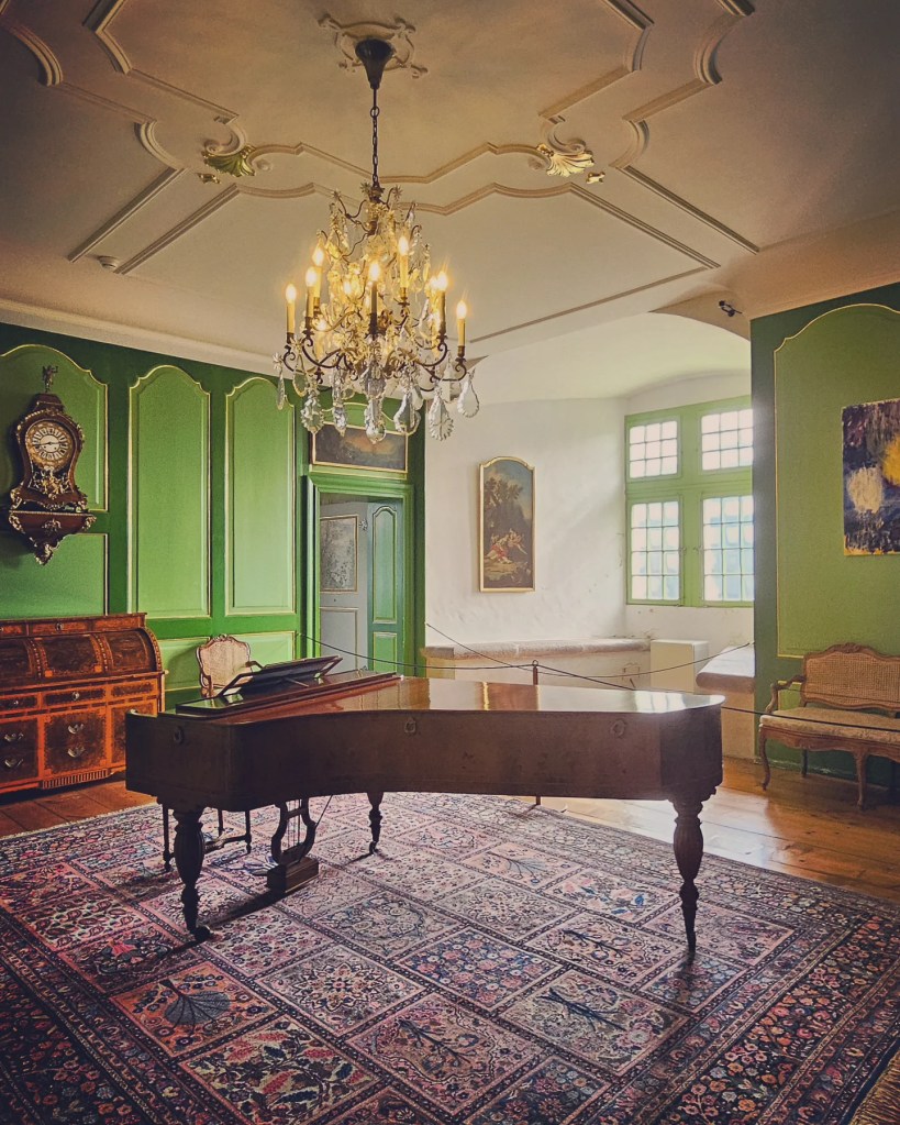 Sun-lit salon painted green, crystal chandelier above a grand piano and Persian rug.