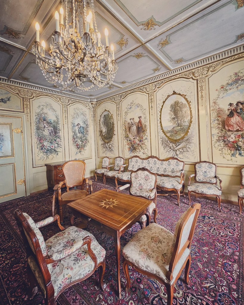 Elegant drawing room in Château de Gruyères with crystal chandelier, floral chairs, painted wall panels and a small wooden game table.