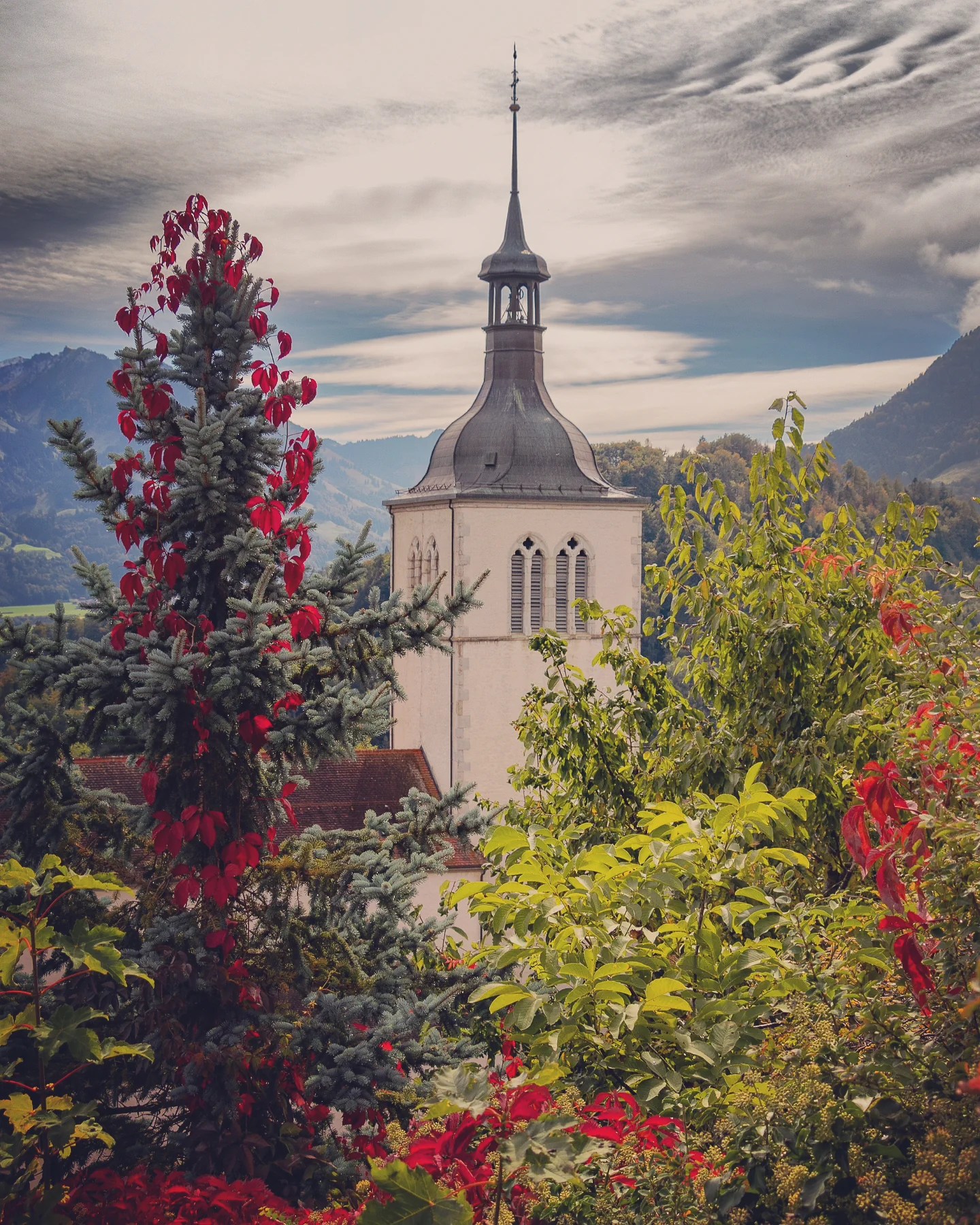 Church tower of Saint-Théodule framed by red vines, evergreens and distant mountains.