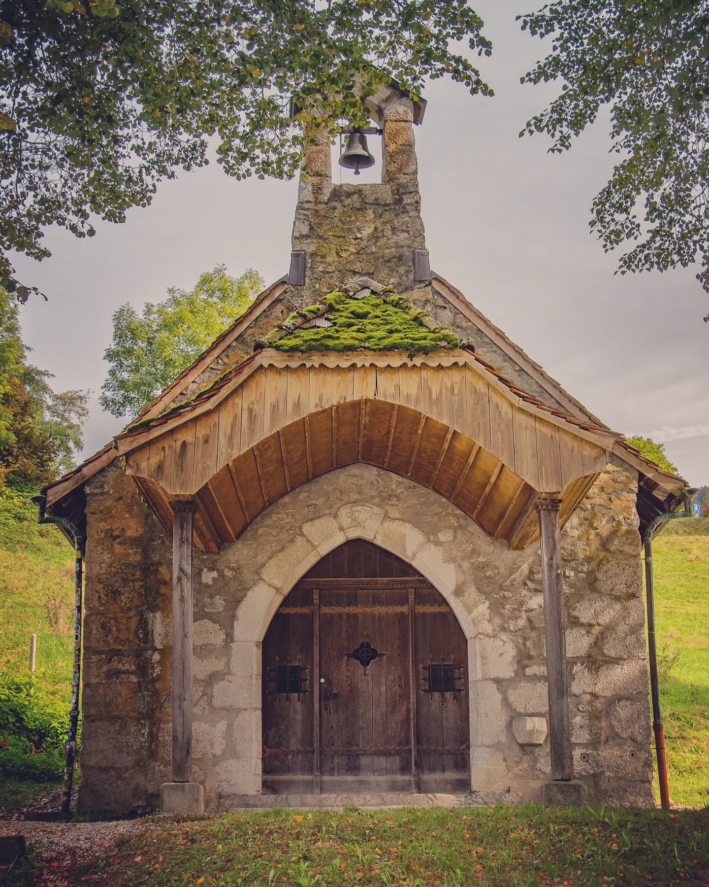 Tiny field chapel with mossy roof tiles, stone bell gable and wooden doors under leafy trees.