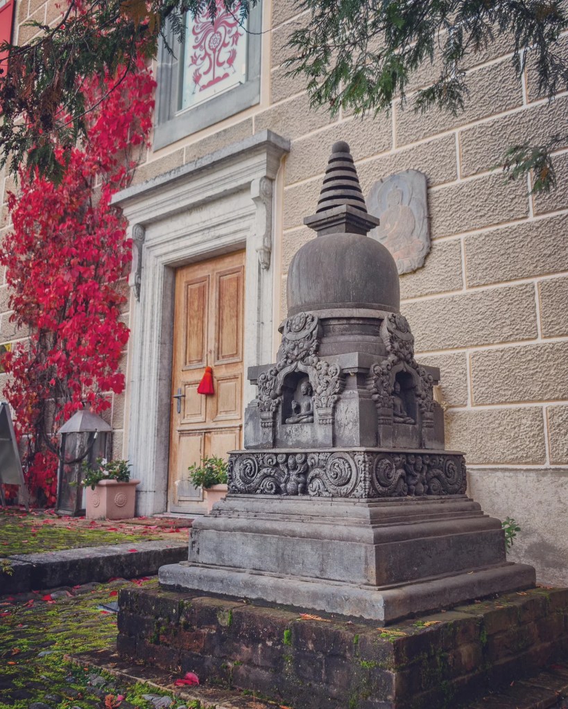 Carved stone stupa outside Gruyères’ Tibet Museum, with red vine creeping up a townhouse wall.