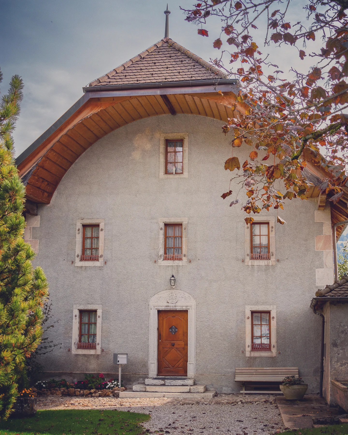 Front view of a traditional Fribourg farmhouse with a high arched roof, stone façade, small wooden bench and autumn leaves in foreground.