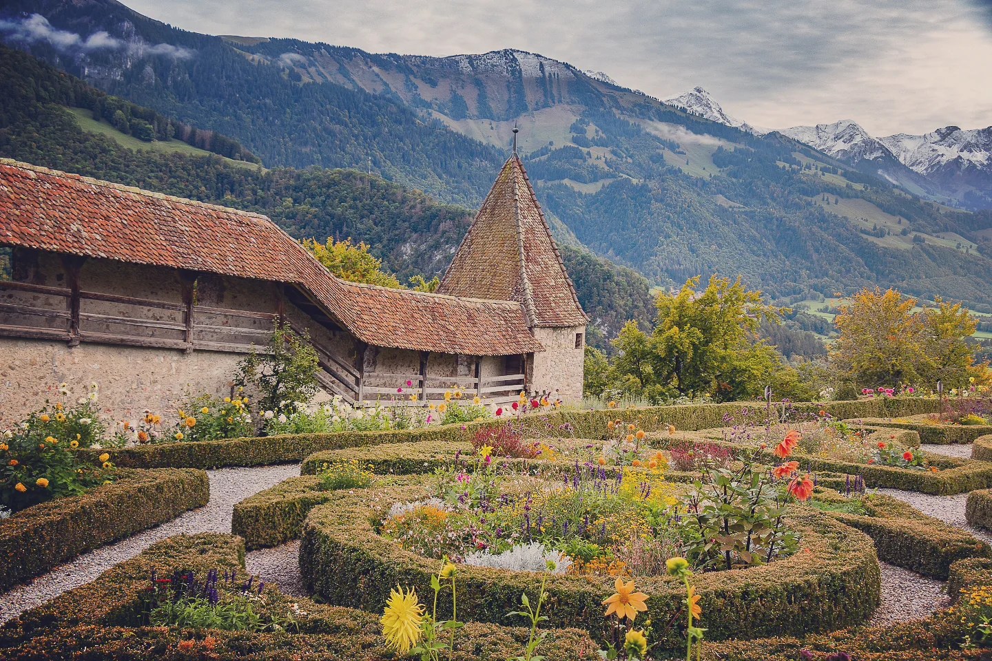 Formal flower beds bordered by boxwood with a stone watch-tower and jagged Pre-Alpine peaks dusted in snow behind.