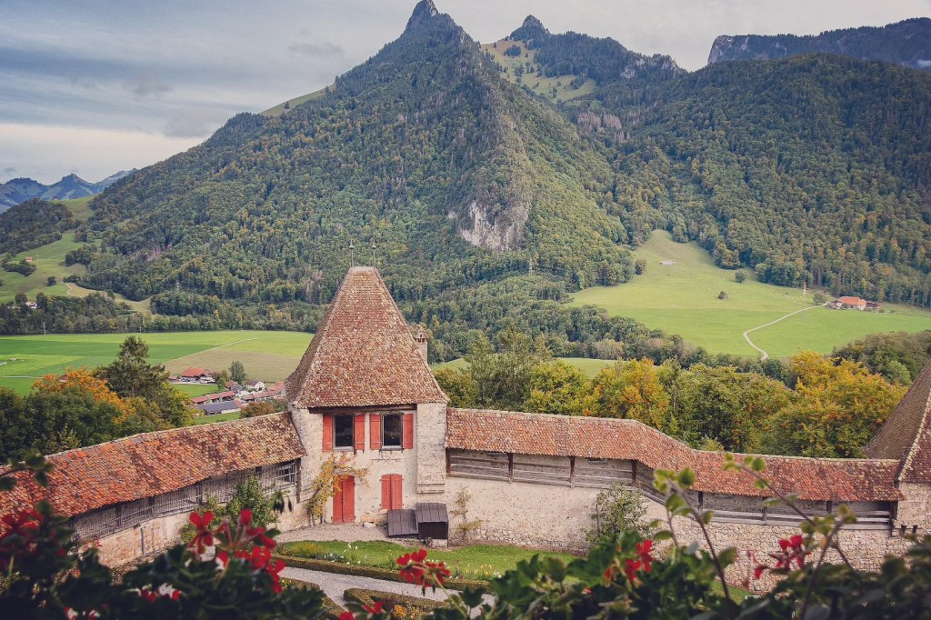 Stone watch-tower with red shutters on Gruyères castle wall; Dent de Broc mountain rises forest-green behind, alpine pastures and farm lanes below.