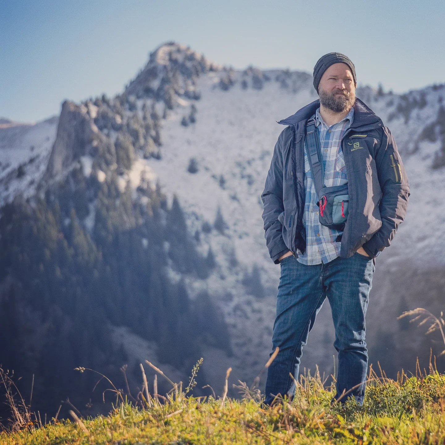 Bearded man in outdoor clothing stands on a grassy ridge with a freshly snowed mountain backdrop.