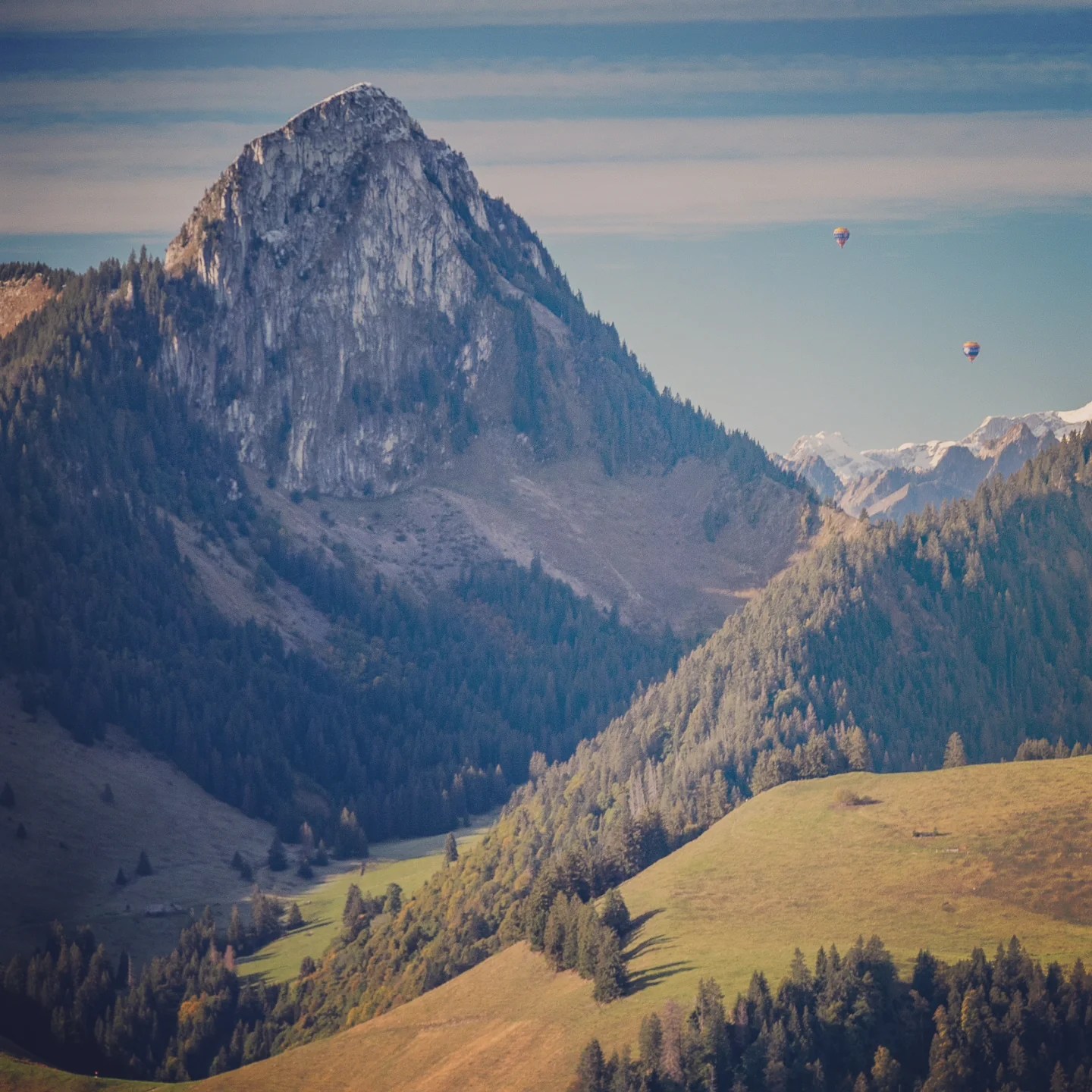Jagged limestone summit rises above wooded slopes; two hot-air balloons drift in the distance.