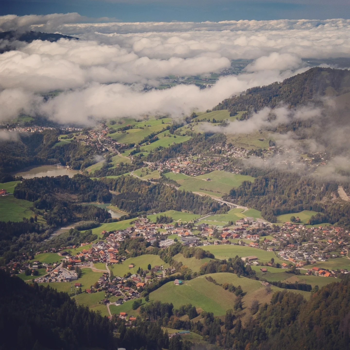 Aerial view of green valleys, villages and river bends partly hidden by a layer of white clouds.