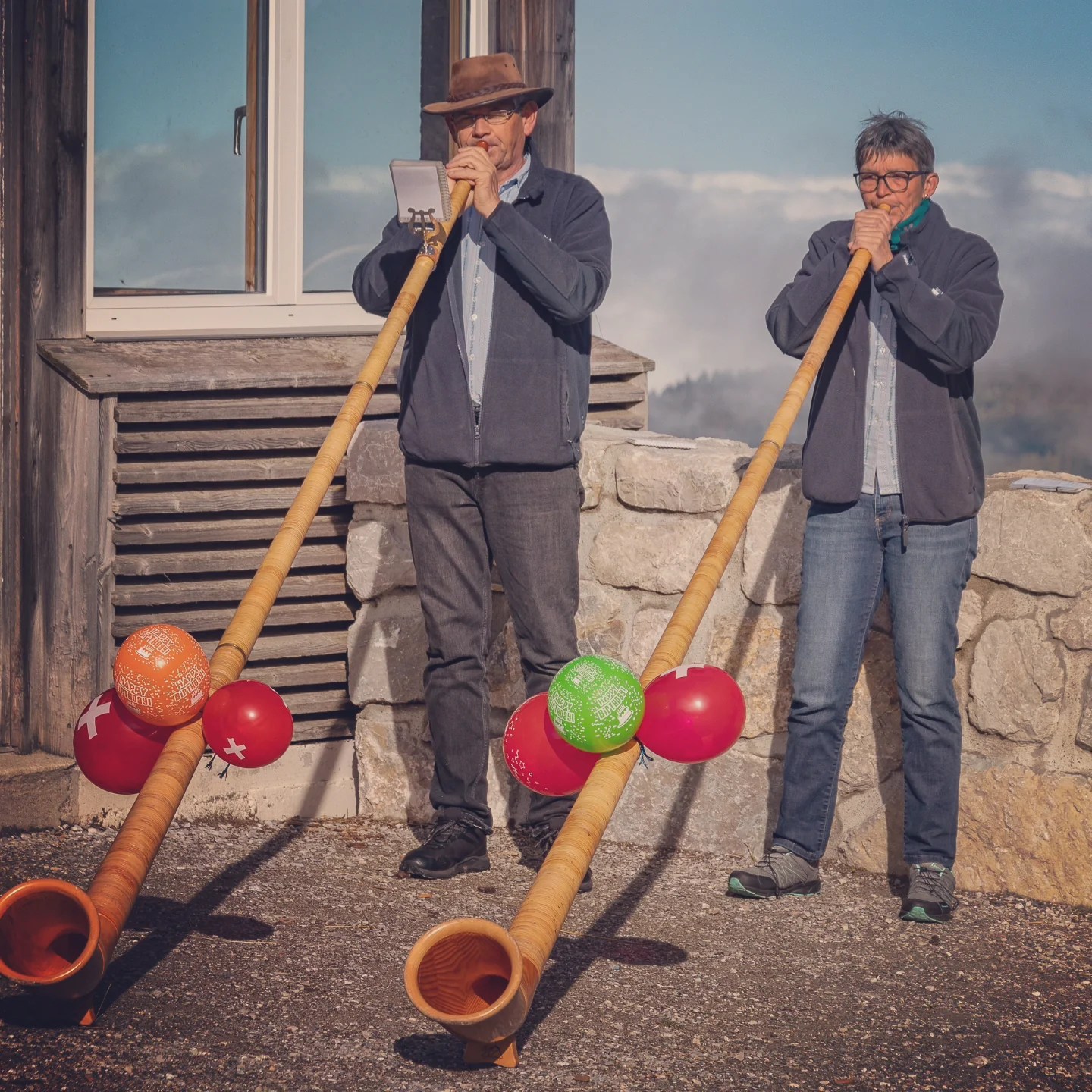 Two musicians play long wooden alphorns decorated with Swiss-flag balloons outside the Charmey mountain gondola building.