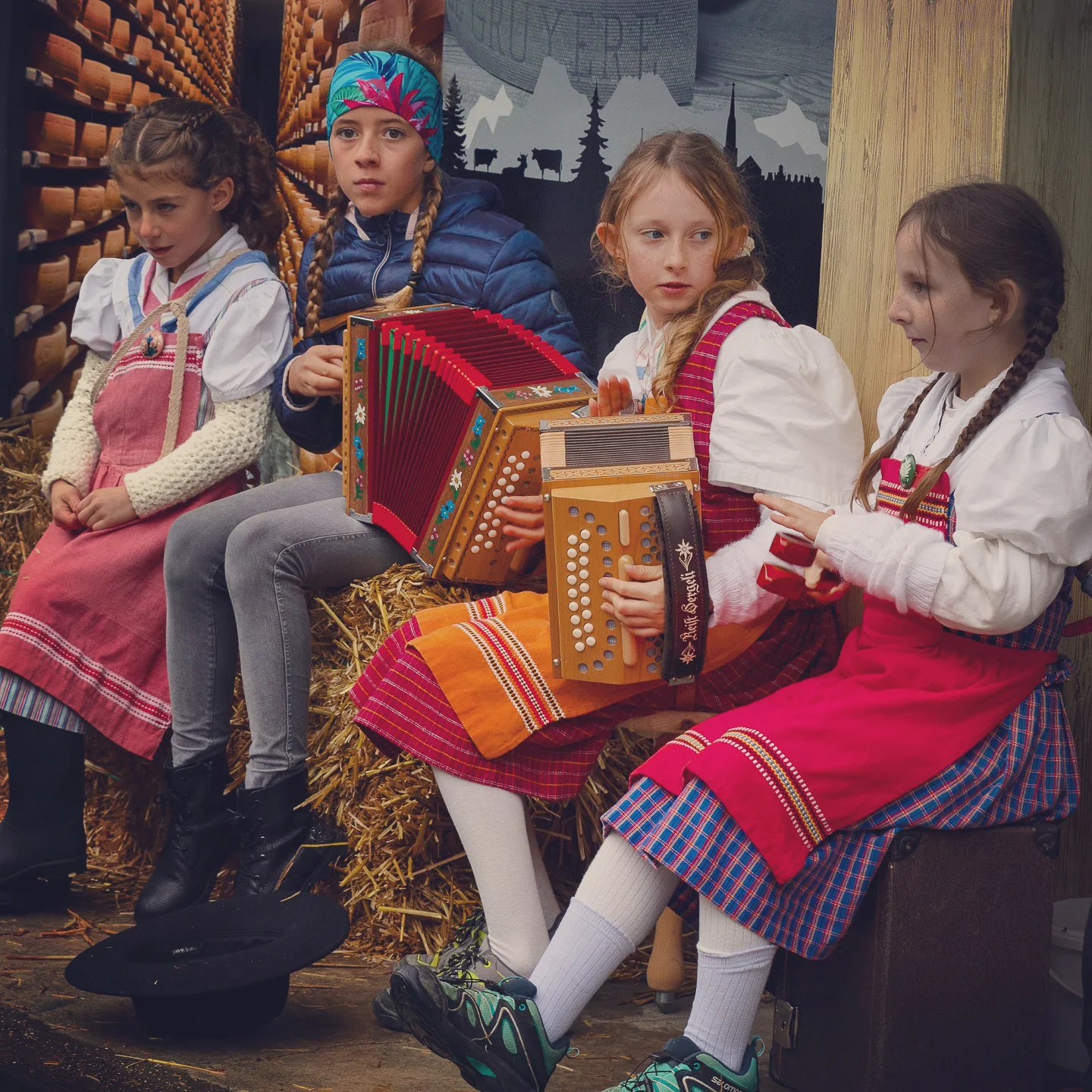 Four girls in traditional dresses sit on straw bales, playing small button accordions and chatting.